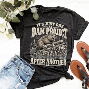 Pode incluir: Uma t-shirt de manga curta cinzento-carvão escuro com um gráfico de um castor numa barragem e o texto IT'S JUST ONE DAM PROJECT AFTER ANOTHER numa fonte branca desgastada, rodeado por uma ilustração de floresta e pequenos pinheiros.