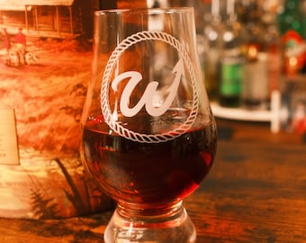 May include: A clear glass whiskey tumbler featuring an etched design of a rope circle surrounding the letter W. The glass is filled with a dark amber liquid and sits next to a cylindrical container decorated with a cabin and forest landscape scene.