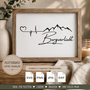 May include: A rectangular wooden frame containing a white print with a black graphic design. The design features a heart, an EKG heartbeat line, a mountain range silhouette, and the German text Bergverliebt in a cursive script font.
