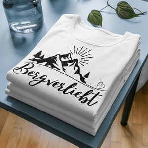 以下が含まれることがあります： 黒い山のグラフィックと「Bergverliebt」というテキストと小さなハートが描かれた白いTシャツ