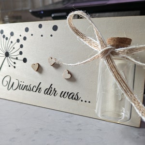 Puede incluir: Un letrero de madera con un diseño de diente de león y el texto "Wunsch dir was..." en negro. Hay una pequeña botella de vidrio con un tapón de corcho y un lazo de cuerda marrón. La botella está atada con un lazo de cuerda marrón. Hay tres pequeños corazones de madera unidos al letrero.