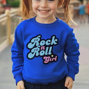 May include: A young girl wearing a blue sweatshirt with the text "Rock 'n' Roll Girl" in a retro font. The sweatshirt has a pink heart and a gold star.