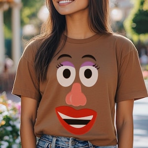 May include: A brown t-shirt with a cartoon face printed on the front. The face has large eyes, a red mouth, and a pink nose.