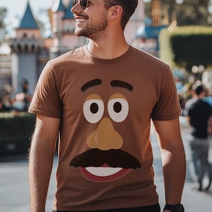 Può includere: Una maglietta marrone con un viso di cartone animato stampato sul davanti. Il viso ha sopracciglia marroni, grandi occhi bianchi con pupille marroni, un naso marrone, un baffo nero e una bocca rossa.