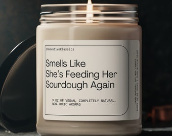 May include: A candle in a clear glass jar with a beige label that reads "Smells Like She's Feeding Her Sourdough Again". The label also says "9 oz of vegan, completely natural, non-toxic aromas".