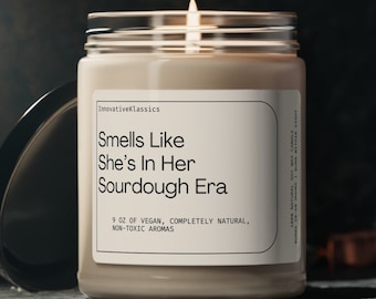 May include: A glass candle jar with a beige label that reads "Smells Like She's In Her Sourdough Era" and "9 oz of vegan, completely natural, non-toxic aromas". The candle is lit and the flame is visible.