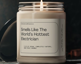 May include: A beige candle in a glass jar with a white label that reads "Smells Like The World's Hottest Electrician". The candle is lit and the flame is visible.
