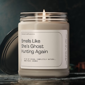 May include: A beige candle with a glass jar and a black lid. The candle is labeled "Smells Like She's Ghost Hunting Again" and "9 oz of Vegan, Completely Natural, Non-Toxic Aromas".