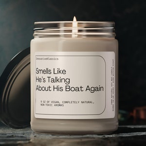 Op de afbeelding: Een beige kaars in een glazen pot met een wit etiket waarop staat "Smells Like He's Talking About His Boat Again". Het etiket vermeldt ook "9 oz Vegan, volledig natuurlijk, niet-giftige aroma's".