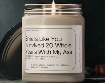 May include: A lit, cream-colored candle in a clear glass jar with a silver lid. The label reads "Smells Like You Survived 20 Whole Years With My Ass." The candle is 9 oz and made of vegan, completely natural, non-toxic aromas.