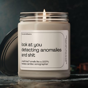May include: A beige candle with a glass jar and a black lid. The candle has a white label with the text "look at you detecting anomalies and shit smell that? smells like a 100% badass cardiac sonographer".