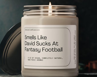 May include: A beige candle with a glass jar and a black lid. The candle has a white label with the text "Smells Like David Sucks At Fantasy Football". The label also says "9 oz of vegan, completely natural, non-toxic aromas".