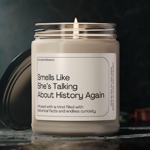 May include: A beige candle with a clear glass jar and a white label that reads "Smells Like She's Talking About History Again. Infused with a mind filled with historical facts and endless curiosity."