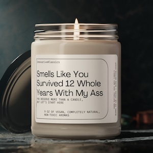 May include: A beige candle with a clear glass jar and a black lid. The candle has a white label with the text "Smells Like You Survived 12 Whole Years With My Ass" and "9 oz of vegan, completely natural, non-toxic aromas".
