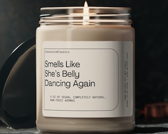 May include: A beige candle with a glass jar and a white label that reads "Smells Like She's Belly Dancing Again". The candle is lit and the flame is visible. The candle is on a dark surface.