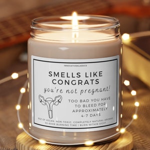 May include: A lit candle in a clear glass jar with a white label that reads "SMELLS LIKE CONGRATS you're not pregnant!" The candle sits on a wooden coaster. A string of lights and a guitar are in the background.