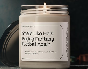 May include: A beige candle with a clear glass jar and a black lid. The candle is labeled "Smells Like He's Playing Fantasy Football Again". The label also states that the candle is 9 oz of vegan, completely natural, non-toxic aromas.
