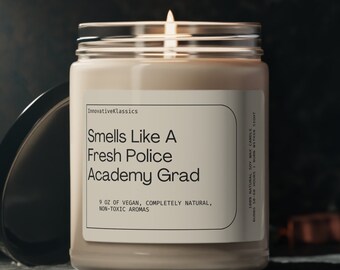 Puede incluir: Una vela beige en un tarro de vidrio con una etiqueta blanca que dice "Smells Like A Fresh Police Academy Grad". La vela está encendida y la llama es visible. La vela está sobre una superficie gris oscura.