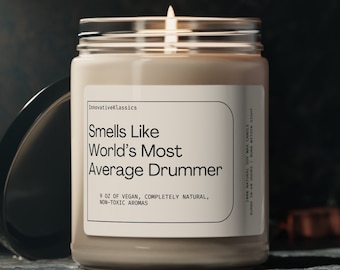 May include: A beige candle in a glass jar with a white label that reads "Smells Like World's Most Average Drummer". The label also says "9 oz of vegan, completely natural, non-toxic aromas".