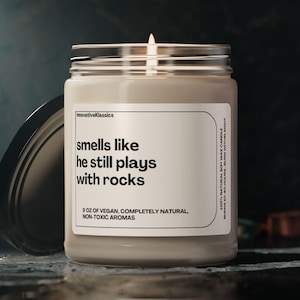 May include: A beige candle with a glass jar and a black lid. The candle has a white label with the text "smells like he still plays with rocks" and "8 oz of vegan, completely natural, non-toxic aromas".