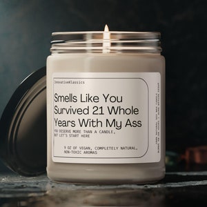 May include: A beige candle with a black lid. The candle label reads "Smells Like You Survived 21 Whole Years With My Ass". The label also says "You deserve more than a candle, but let's start here." and "9 oz of vegan, completely natural, non-toxic aromas".