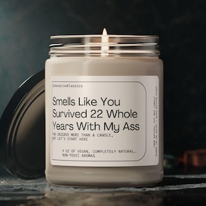 May include: A beige candle with a black lid. The candle has a white label with the text "Smells Like You Survived 22 Whole Years With My Ass" and "5 oz of vegan, completely natural, non-toxic aromas".