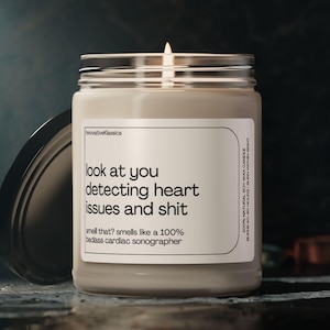 May include: A beige candle with a glass jar and a black lid. The candle has a white label with the text "look at you detecting heart issues and shit smell that? smells like a 100% badass cardiac sonographer".