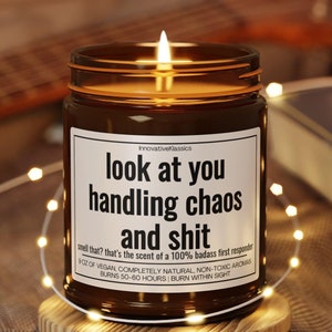Puede incluir: Una vela de vidrio marr&oacute;n con una etiqueta blanca que dice "look at you handling chaos and shit". La vela est&aacute; encendida y la llama es visible. El texto en la etiqueta tambi&eacute;n dice "that's the scent of a 100% badass first responder".
