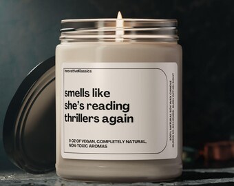 May include: A beige candle with a glass jar and a black lid. The candle has a white label with the text "smells like she's reading thrillers again" and "8 oz of vegan, completely natural, non-toxic aromas".