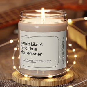 May include: A brown candle in a glass jar with a white label that reads "Smells Like A First Time Homeowner 9 oz of Vegan, Completely Natural, Non-Toxic Aromas". The candle is lit and sitting on a wooden coaster.
