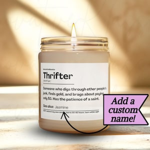 May include: A lit candle in a glass jar with a white label. The label reads "Thrifter" with a definition. A pink banner on the right says "Add a custom name!" The candle is on a light-colored surface.