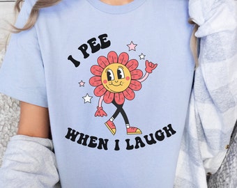 Op de afbeelding: Een lichtblauw T-shirt met een cartoon-bloemdessin. De bloem heeft een glimlachend gezicht en draagt schoenen. De tekst "I pee when I laugh" is op het T-shirt gedrukt.