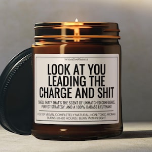 May include: A brown glass candle jar with a black lid. The candle jar has a white label with the text "LOOK AT YOU LEADING THE CHARGE AND SHIT" and the text "SMELL THAT? THAT'S THE SCENT OF UNMATCHED CONFIDENCE, PERFECT STRATEGY, AND A 100% BADASS LIEUTENANT". The label also includes the text "9OZ OF VEGAN, COMPLETELY NATURAL, NON-TOXIC AROMAS BURNS 50-60 HOURS BURN WITHIN SIGHT".