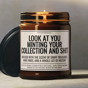 Puede incluir: Un tarro de vela de vidrio marr&oacute;n con una tapa negra. La vela est&aacute; etiquetada "LOOK AT YOU MINTING YOUR COLLECTION AND SHIT" y dice "INFUSED WITH THE SCENT OF SHINY TREASURES, RARE FINDS, AND A WHOLE LOT OF HISTORY." La vela es de 8 oz de aromas veganos, completamente naturales y no t&oacute;xicos y arde durante 50-60 horas.