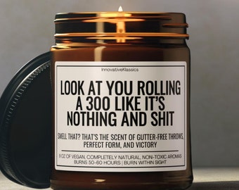 May include: A brown glass candle jar with a black lid. The candle has a white label with the text "LOOK AT YOU ROLLING A 300 LIKE IT'S NOTHING AND SHIT" and the text "SWELL THAT? THAT'S THE SCENT OF GUTTER-FREE THROWS, PERFECT FORM, AND VICTORY". The label also includes the text "8 OZ OF VEGAN, COMPLETELY NATURAL, NON-TOXIC AROMAS BURNS 50-60 HOURS | BURN WITHIN SIGHT".