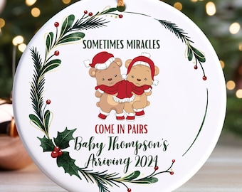 May include: White ceramic Christmas ornament with a green wreath design. The ornament features two cartoon bears wearing Santa hats and the text "Sometimes miracles come in pairs Baby Thompson's Arriving 2024".