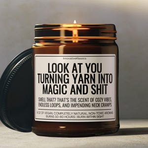 May include: A brown glass candle jar with a black lid. The candle has a white label with the text "LOOK AT YOU TURNING YARN INTO MAGIC AND SHIT" and the description "SMELL THAT? THAT'S THE SCENT OF COZY VIBES, ENDLESS LOOPS, AND IMPENDING NECK CRAMPS 8 OZ OF VEGAN, COMPLETELY NATURAL, NON-TOXIC AROMAS BURNS 50-60 HOURS | BURN WITHIN SIGHT".