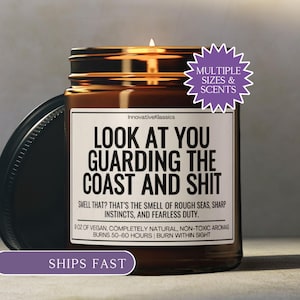 May include: A brown glass candle jar with a black lid, featuring a white label with black text that reads "LOOK AT YOU GUARDING THE COAST AND SHIT." The candle is labeled as 9 oz of vegan, non-toxic aromas and burns for 50-60 hours.