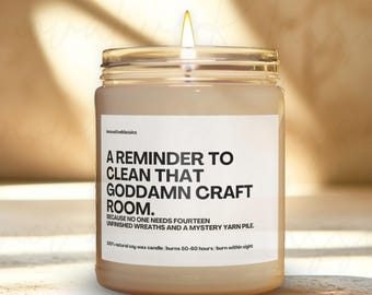 May include: A lit soy wax candle in a clear glass jar. The label reads "A REMINDER TO CLEAN THAT GODDAMN CRAFT ROOM." The text continues, "BECAUSE NO ONE NEEDS FOURTEEN UNFINISHED WREATHS AND A MYSTERY YARN PILE."