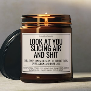 May include: A lit brown glass jar candle with a black lid. The candle has a white label with black text that reads "LOOK AT YOU SLICING AIR AND SHIT". The text below reads "SMELL THAT? THAT'S THE SCENT OF PERFECT TIMING, SWIFT ACTION, AND PURE SKILL".