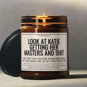 May include: A brown glass candle jar with a black lid. The candle jar has a white label with the text "LOOK AT KATIE GETTING HER MASTERS AND SHIT" and the text "SMELL THAT? THAT'S THE SCENT OF HARD WORK PAYING OFF, A 100% BADASS, AND I AM SO F*CKING PROUD OF YOU". The label also includes the text "9 OZ OF VEGAN, COMPLETELY NATURAL, NON-TOXIC AROMAS BURNS 50-60 HOURS | BURN WITHIN SIGHT".