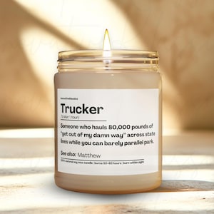 May include: A lit candle in a clear glass jar with a white label. The label reads "Trucker" with a definition and the text "See also: Matthew". The candle is on a neutral surface.