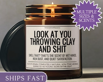 May include: A lit brown glass jar candle with a black lid. The label reads "LOOK AT YOU THROWING CLAY AND SHIT." The candle is 9 oz of vegan, non-toxic aromas and burns for 50-60 hours. A purple sticker says "MULTIPLE SIZES & SCENTS."