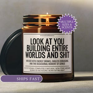 May include: A brown glass jar candle with a black lid, featuring the text "LOOK AT YOU BUILDING ENTIRE WORLDS AND SHIT." The label also includes details about the candle's vegan, natural, and non-toxic composition. The candle burns for 50-60 hours.