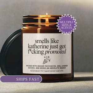 May include: A brown glass candle jar with a black lid and a white label that reads: smells like katherine just got f*cking promoted, infused with bigger paychecks, well-earned success, and being an absolute badass. The candle has a 9 oz weight.