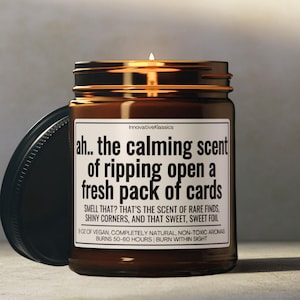 May include: A lit brown glass jar candle with a black lid. The label reads "ah.. the calming scent of ripping open a fresh pack of cards." The text below describes the scent as rare finds, shiny corners, and sweet foil. The candle burns for 50-60 hours.