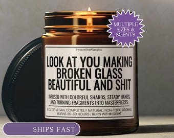 Può includere: Una candela in barattolo di vetro marrone con coperchio nero e etichetta bianca con il testo: LOOK AT YOU MAKING BROKEN GLASS BEAUTIFUL AND SHIT. INFUSED WITH COLORFUL SHARDS, STEADY HANDS, AND TURNING FRAGMENTS INTO MASTERPIECES. 255G di aromi vegani.