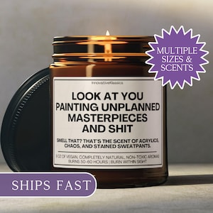 May include: A lit candle in a brown glass jar with a black lid. The label reads "LOOK AT YOU PAINTING UNPLANNED MASTERPIECES AND SHIT." The text also mentions the scent of acrylics, chaos, and stained sweatpants. A purple sticker says "MULTIPLE SIZES & SCENTS."