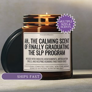 May include: A lit brown glass jar candle with a black lid. The label reads "AH.. THE CALMING SCENT OF FINALLY GRADUATING THE SLP PROGRAM." The candle is infused with natural aromas and burns for 50-60 hours. A purple starburst sticker says "MULTIPLE SIZES & SCENTS."