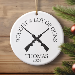 May include: White ceramic Christmas ornament with black text that reads "Bought a lot of guns" with two crossed rifles and the name "Thomas" and the year "2024" below.